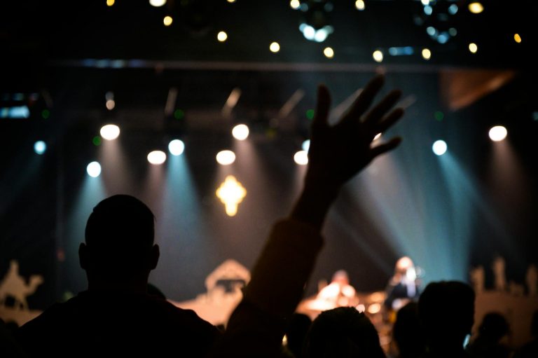 Silhouetted audience raising hands at a vibrant music concert under colorful stage lights.